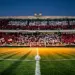 Floodlit football stadium at dusk with crimson and white stand sections, ball on centre circle under spotlight, shadows across pitch.