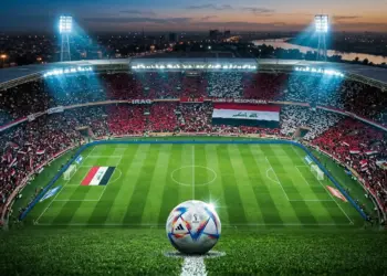 Floodlit stadium pitch at night with Iraqi flag-coloured stands and football on penalty spot.