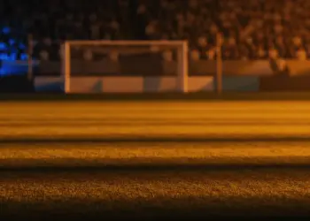Floodlit stadium at golden-hour with packed stands and dramatic shadow patterns under warm and cool atmospheric lighting.