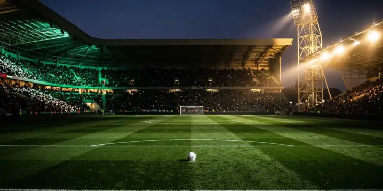 Floodlit stadium pitch at dusk with emerald and white stand sections, centre-spot ball, dramatic shadow play.