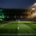 Floodlit stadium pitch at dusk with emerald and white stand sections, centre-spot ball, dramatic shadow play.