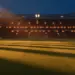 Illuminated stadium pitch at golden hour, dramatic shadows across empty field, packed stands blurred in background.