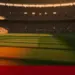 Empty floodlit stadium pitch at dusk with golden lighting and packed stands, no players visible.