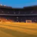 Floodlit stadium pitch at golden hour with shadows, packed stands blurred in background, warm orange and cool blue atmospheric tones.