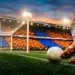 Floodlit stadium pitch with orange and blue stand sections, football boot striking ball with goal net in background, golden evening light and dramatic shadows.
