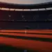 Floodlit stadium pitch with dramatic shadows, packed stands, and golden-hour lighting; no players visible.