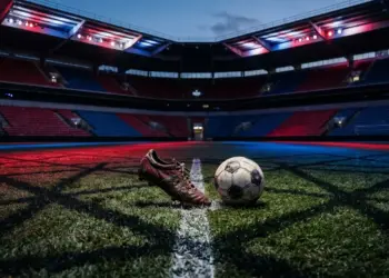 Floodlit stadium pitch at dusk with dramatic red and blue lighting, empty stands, boot striking ball at centre spot, geometric shadows.