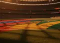 Floodlit stadium pitch with red-blue and gold-green flag colours reflecting on grass at dusk, packed stands in soft focus background.