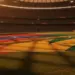 Floodlit stadium pitch with red-blue and gold-green flag colours reflecting on grass at dusk, packed stands in soft focus background.