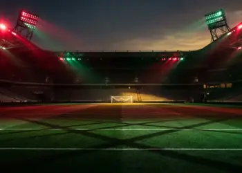 Floodlit stadium pitch in red and green lighting with geometric shadows and billowing goal net.