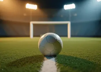 Evening-lit stadium pitch with match ball at penalty spot, golden floodlights casting long shadows, green and blue colour palette.