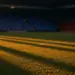 Floodlit stadium pitch at dusk with dramatic shadows and packed stands in evening light, no people visible.
