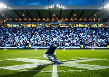 Floodlit stadium with Scotland flag colours, boot on centre pitch, supporter sections in saltire blue and white.