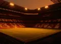 Floodlit stadium with golden-hour lighting, red and yellow colour palette, packed stands and dramatic shadow patterns.