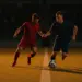 Two footballers in red and navy jerseys contest the ball under floodlights on a stadium pitch with dramatic shadow patterns.