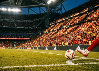 Floodlit football pitch at dusk with Spanish-coloured stand sections and ball in motion during evening match.