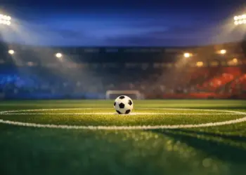 Floodlit football pitch at dusk with match ball on penalty spot, dramatic blue and amber stadium lighting.