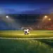 Floodlit football pitch at dusk with match ball on penalty spot, dramatic blue and amber stadium lighting.