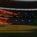 Empty floodlit stadium at dusk with dramatic shadows and packed stands, red-white and green-white national lighting tones.