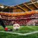 Professional footballer mid-stride with ball on floodlit pitch, Swiss flag colours in crowded stadium stands.
