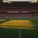 Floodlit football stadium pitch at dusk with shadow patterns, empty field, crowd blur in background.