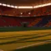 Illuminated stadium pitch at sunset with packed stands, dramatic colour-graded lighting emphasizing red and orange tones.