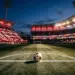 Floodlit stadium pitch with red and white bunting, ball on centre circle, evening light and dramatic shadows.