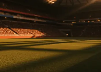 Floodlit stadium at dusk with packed stands and dramatic shadows across empty pitch, golden-hour kick-off atmosphere.