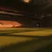 Floodlit stadium at dusk with packed stands and dramatic shadows across empty pitch, golden-hour kick-off atmosphere.