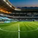 Floodlit stadium pitch at golden hour with white and blue national colours in stand bunting, football on centre circle.
