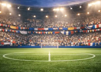 Floodlit stadium with bunting and national-flag colours draped across packed stands, empty pitch bathed in evening spotlight.