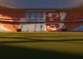Empty floodlit football stadium at dusk with Argentina blue-white and Switzerland red-white coloured stands under dramatic lighting.
