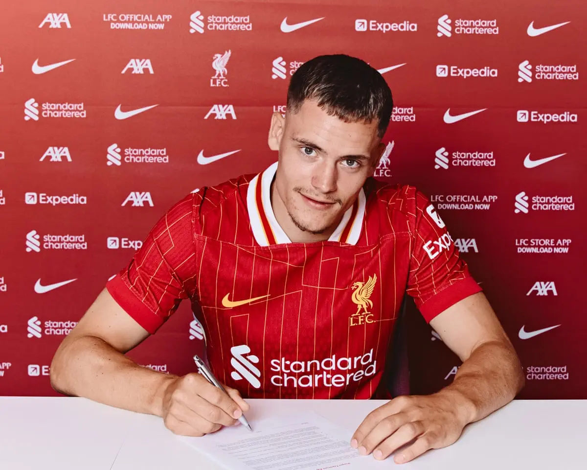 Florian Wirtz wearing a Liverpool FC home kit and signing a contract in front of a club sponsor backdrop