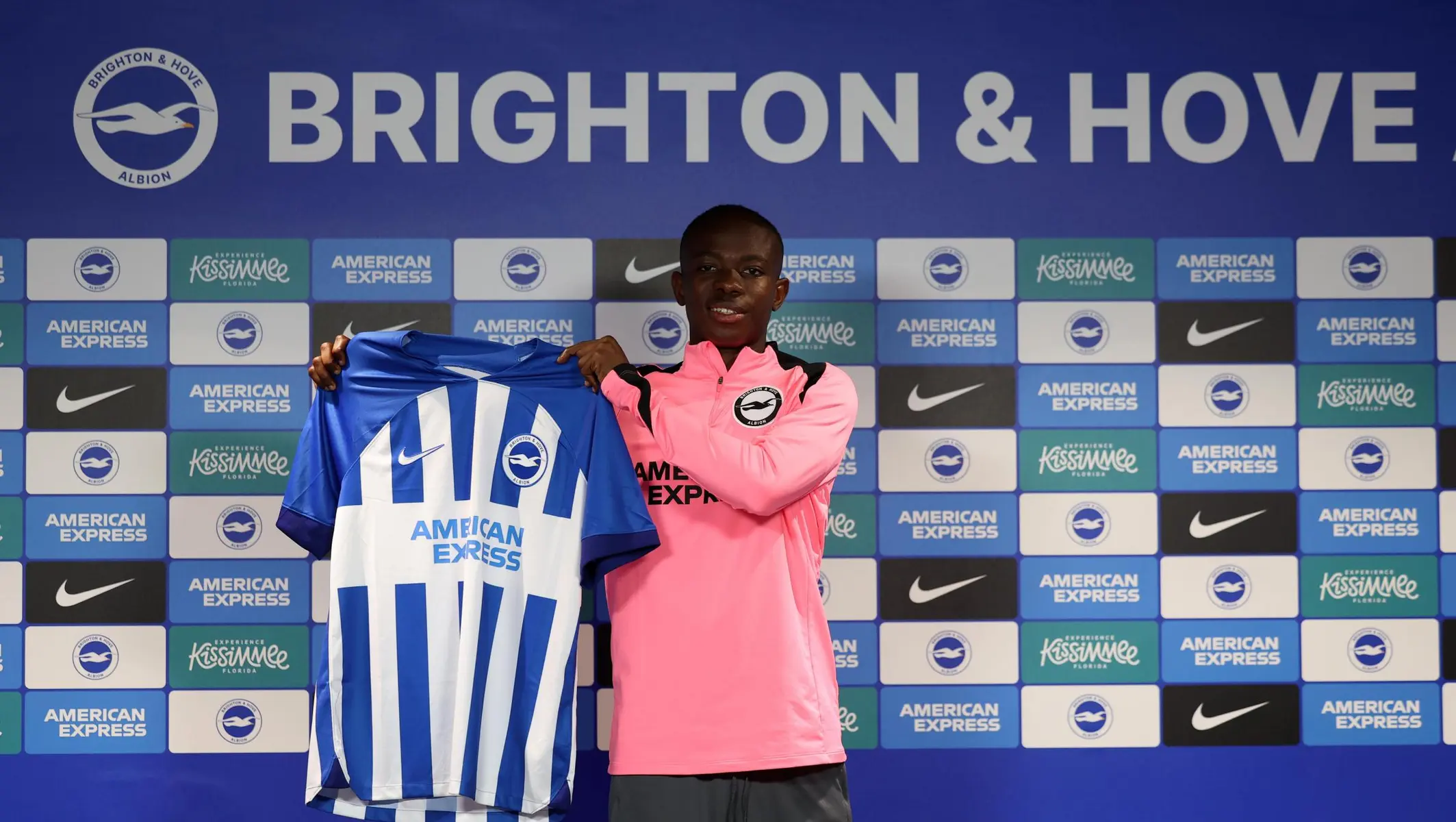 Malick Yalcouyé holding a Brighton & Hove Albion home jersey in front of a club branded backdrop