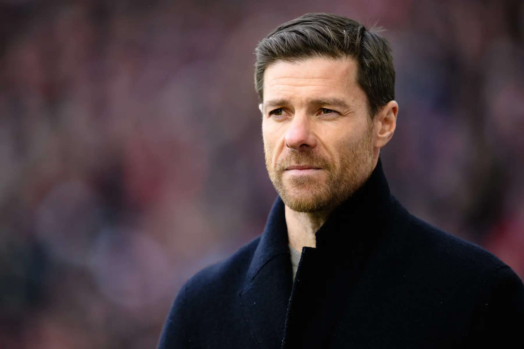 Close-up portrait of Xabi Alonso wearing a dark navy coat against a blurred stadium background