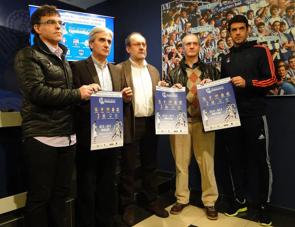 Five men standing together, holding posters for a Real Sociedad youth football program.