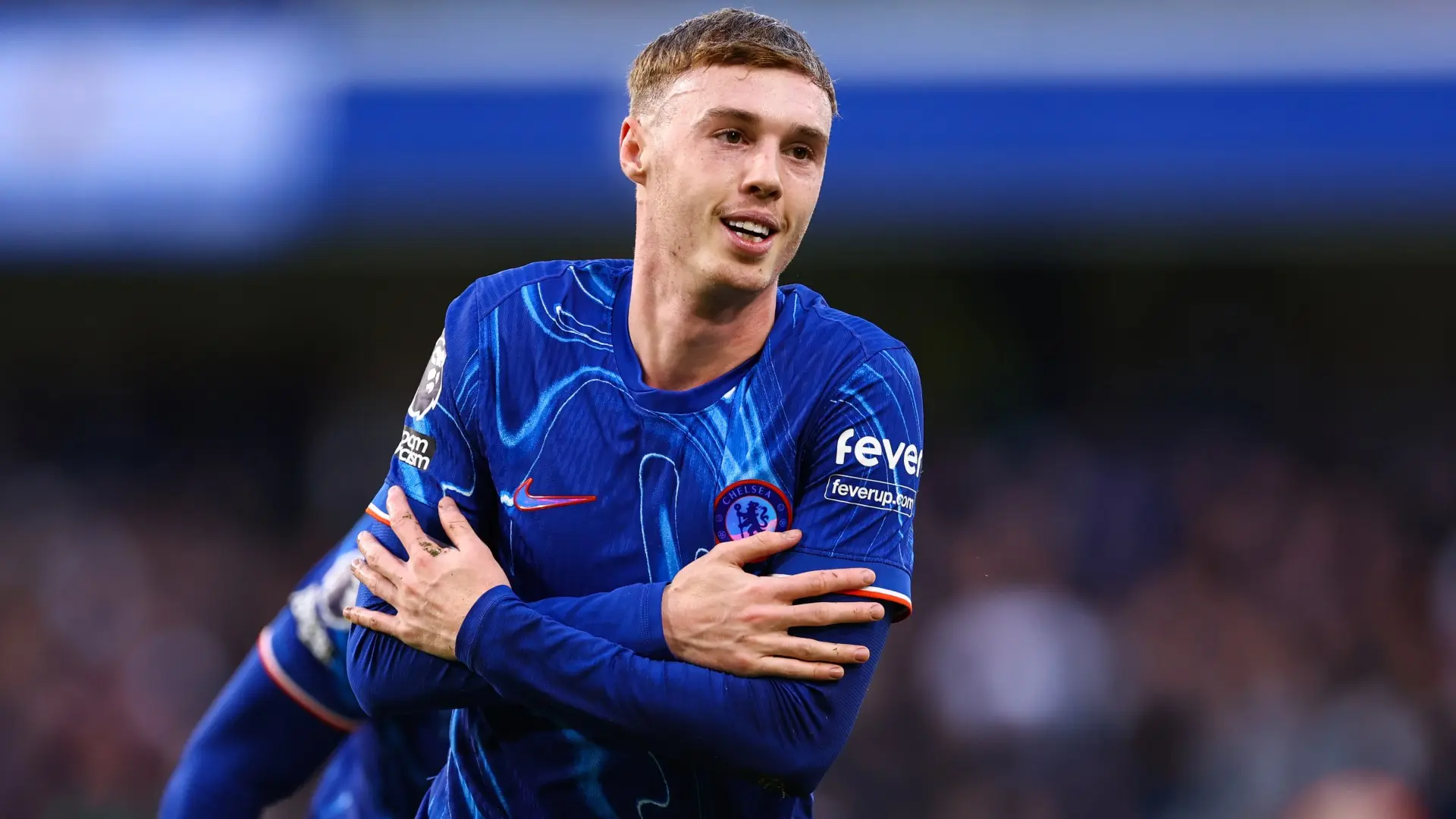 Cole Palmer in a blue Chelsea FC kit performing his signature celebration by crossing his arms over his chest
