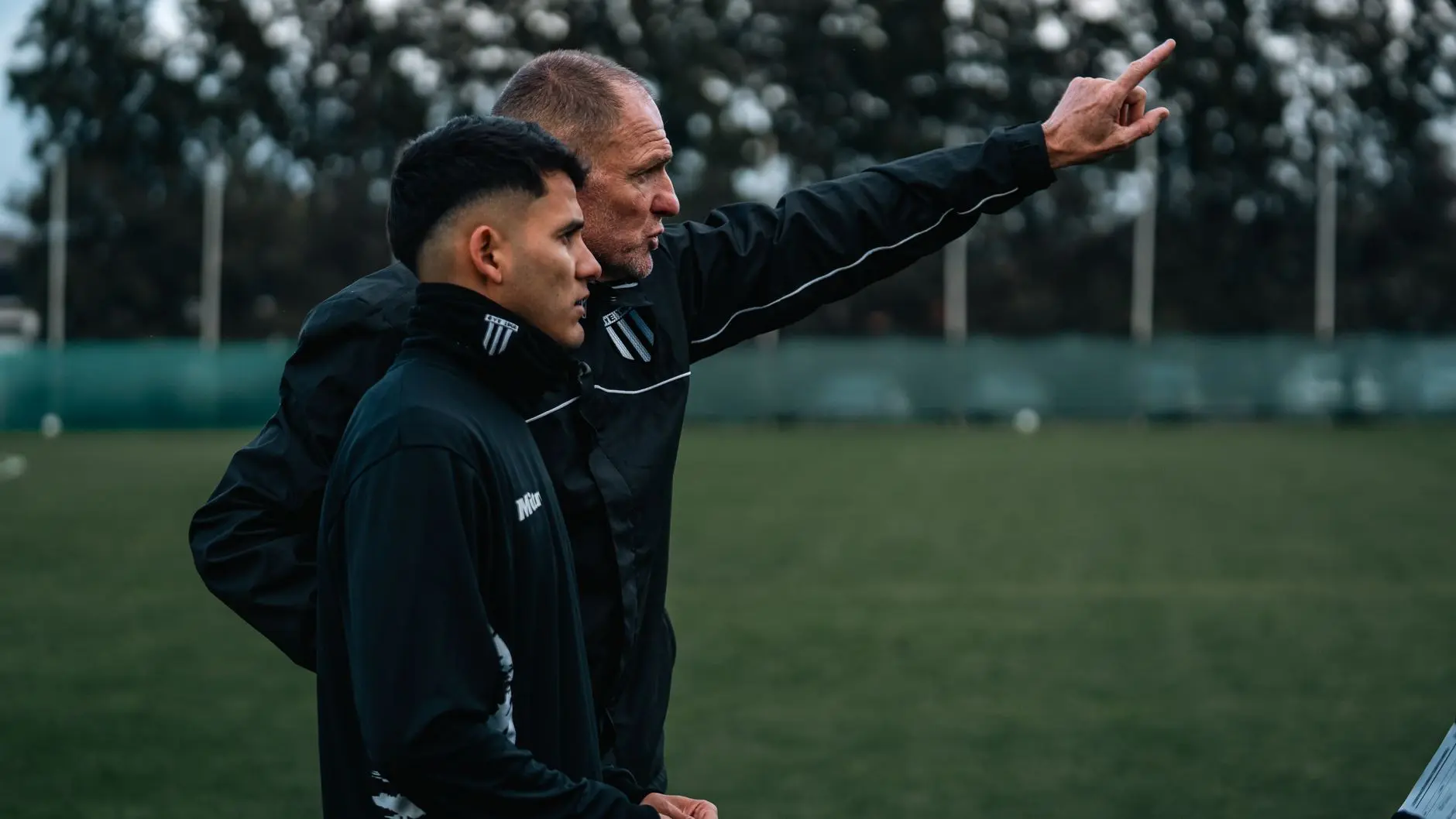 Mauro Lustrinelli in black coaching gear pointing across a green soccer field while talking to a player